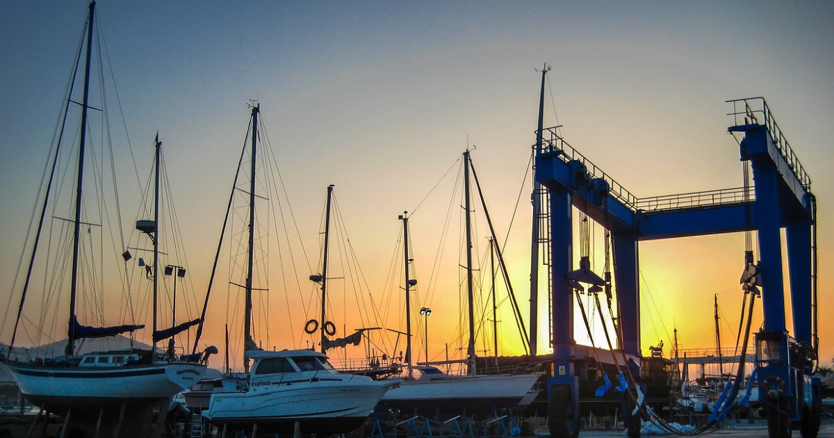 A cluster of boats in a harbour next to a gantry crane with a sunset sky as a backdrop.